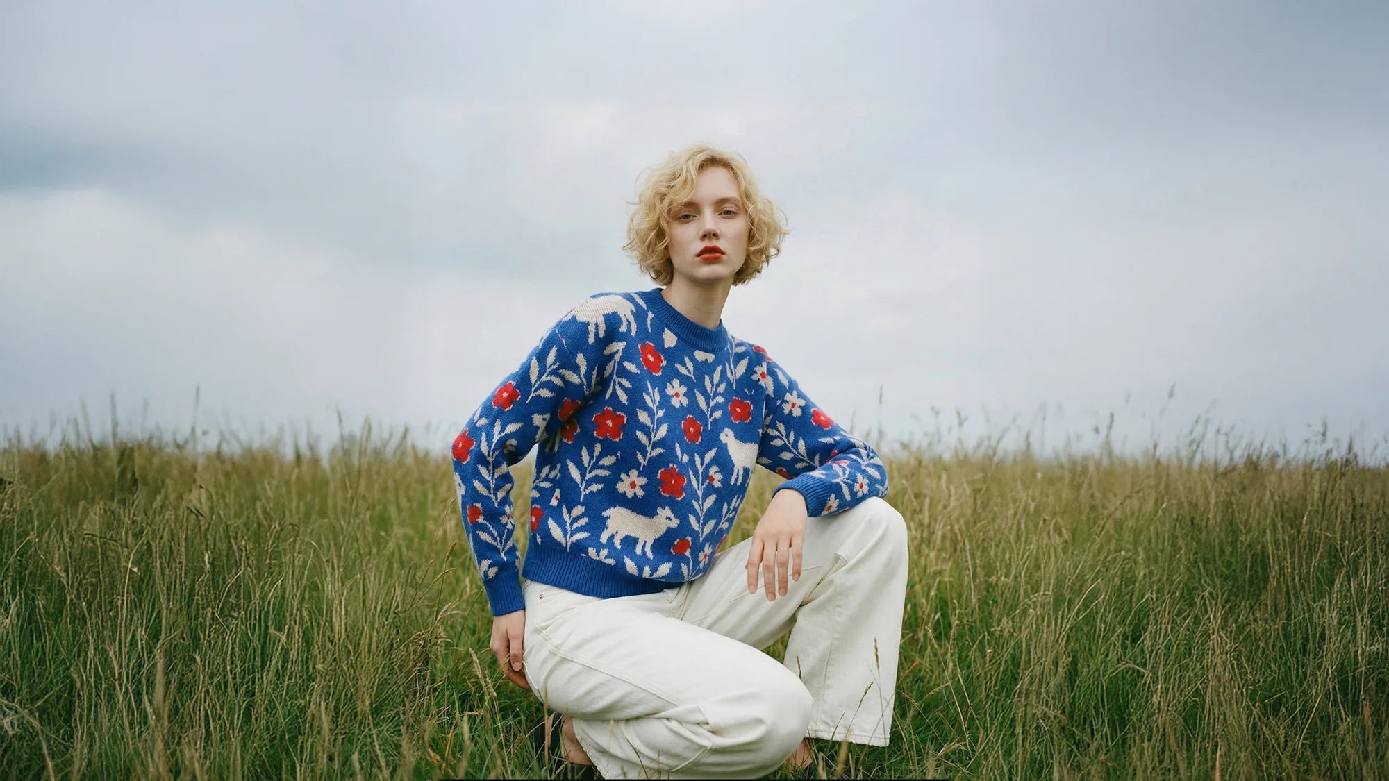 Woman kneeling in tall grass beneath a cloudy sky, wearing a blue floral knit jumper with white animal motifs.
