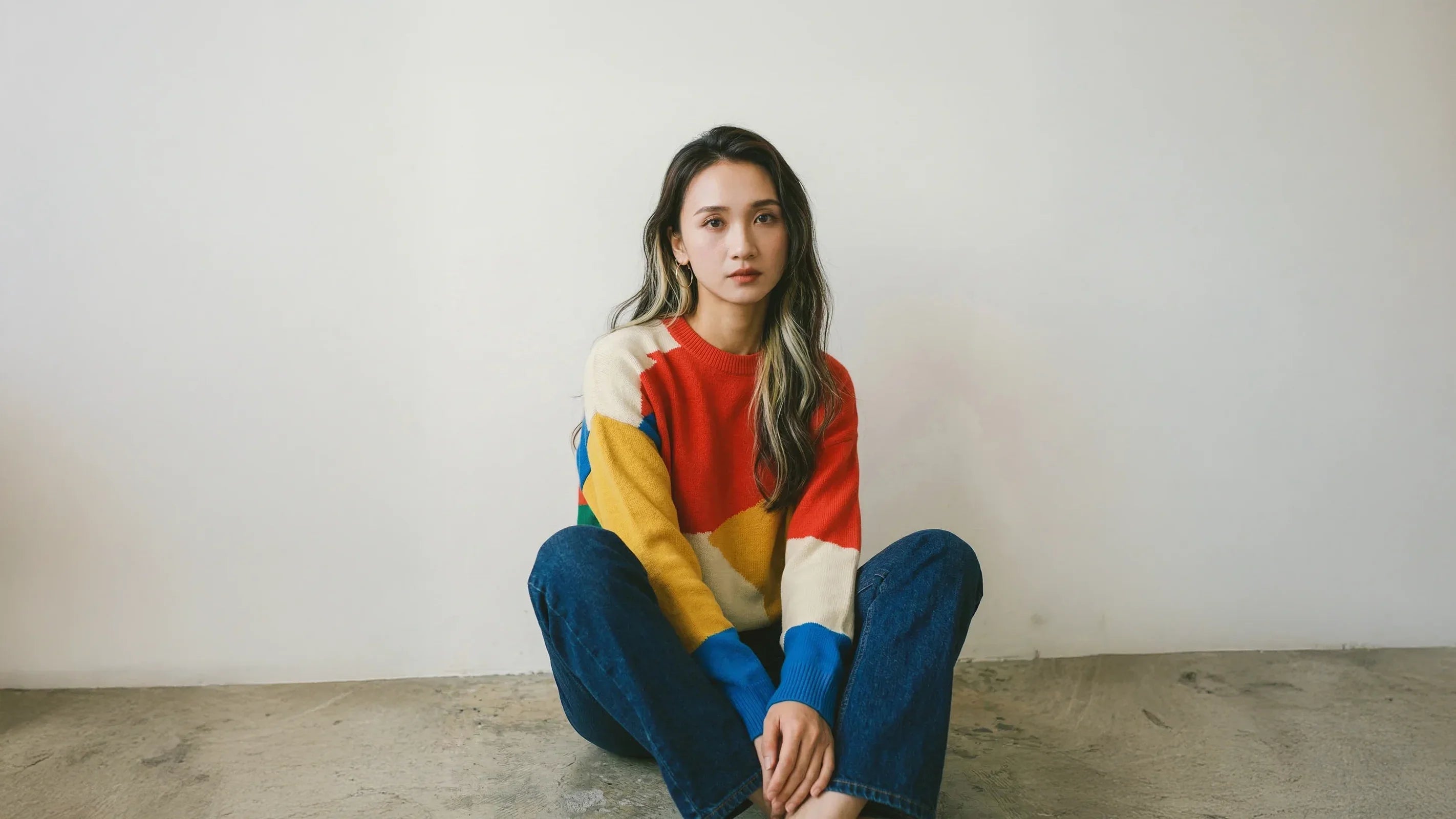 Model seated against a pale wall wearing a colourful graphic jumper with red, yellow, blue and cream panels.