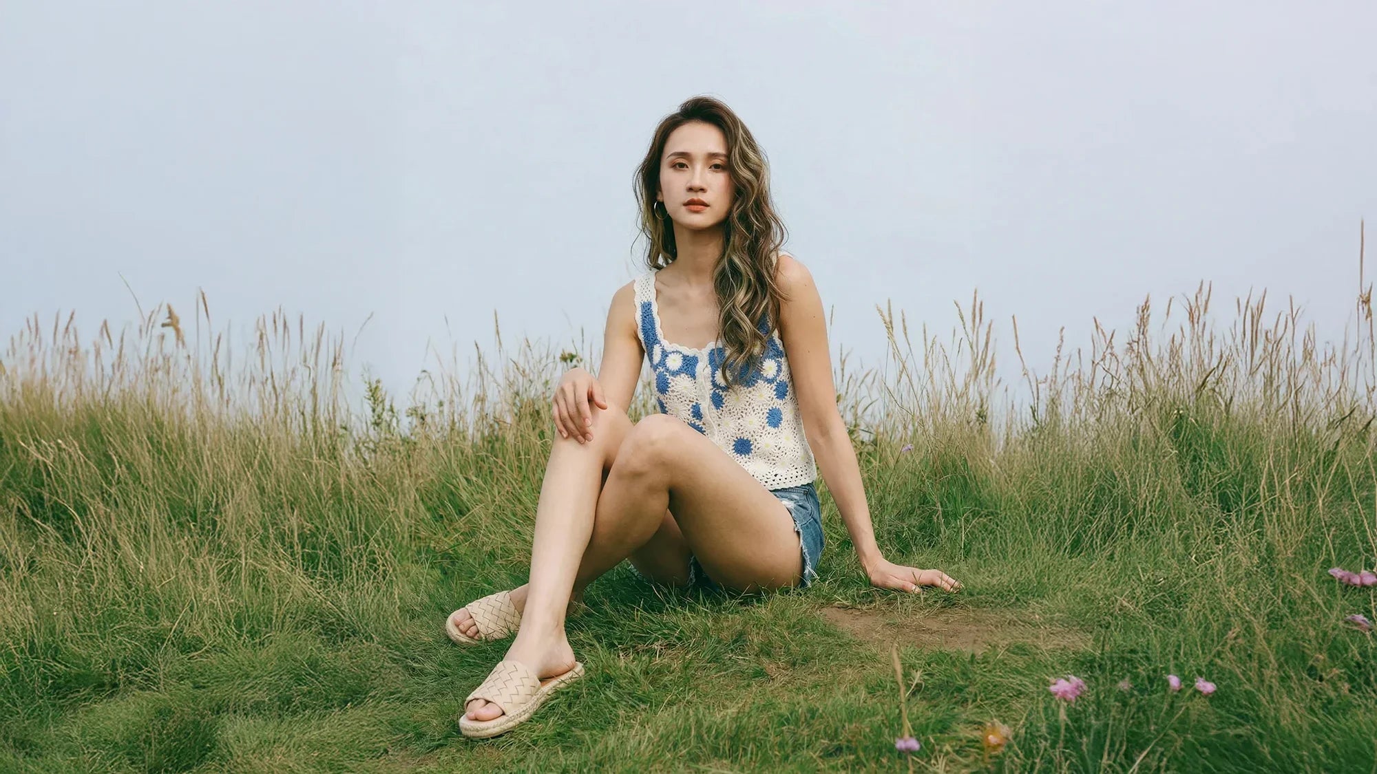 Woman sitting in tall grass wearing a blue and cream crochet top and denim shorts