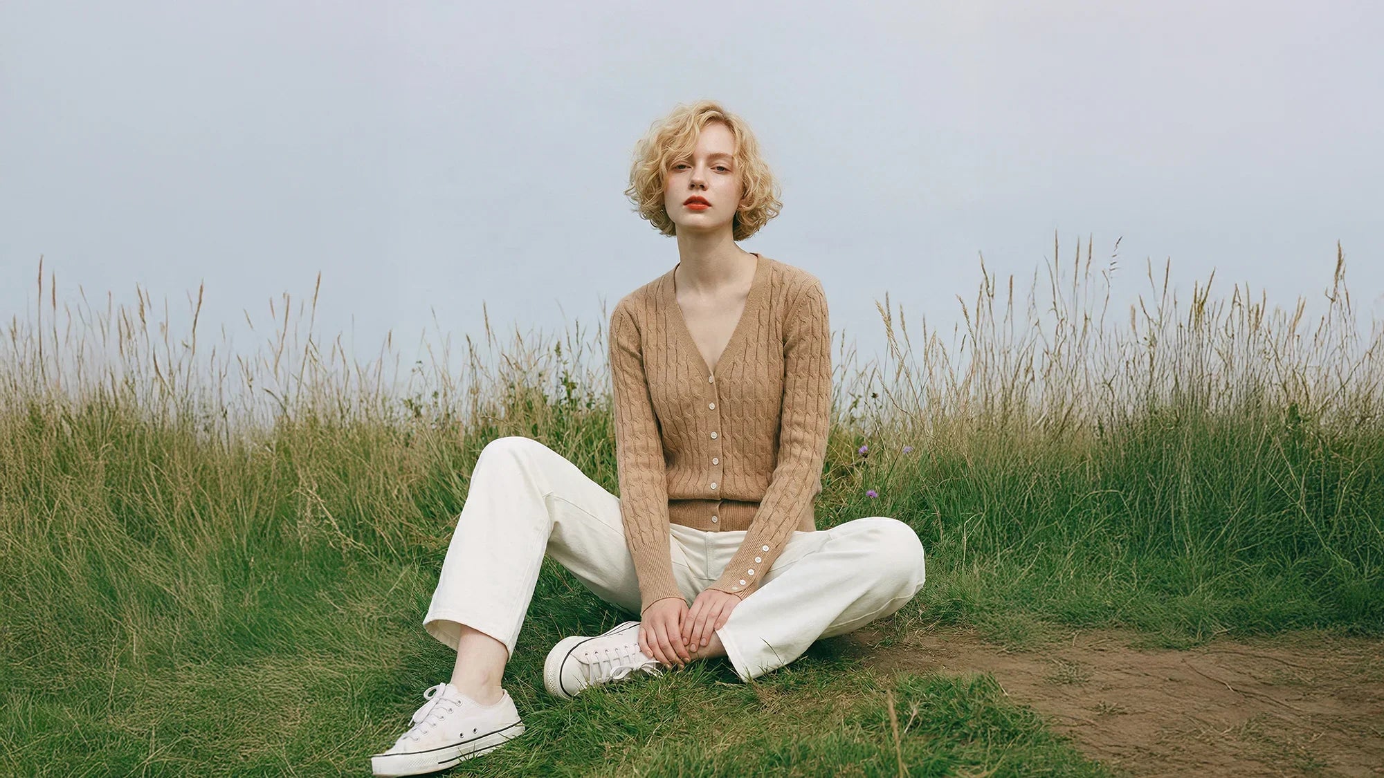Short-haired woman sitting in grass wearing a beige knit cardigan and cream trousers