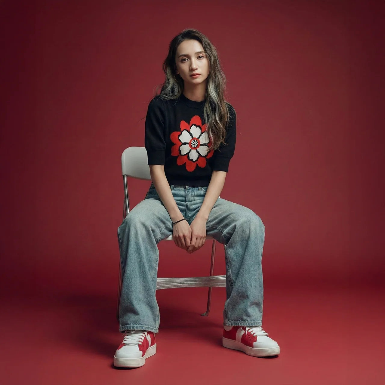 Woman seated on a folding chair against a deep red studio backdrop, wearing a black short-sleeve knit top with a large red and white flower motif, light blue jeans, and red trainers.