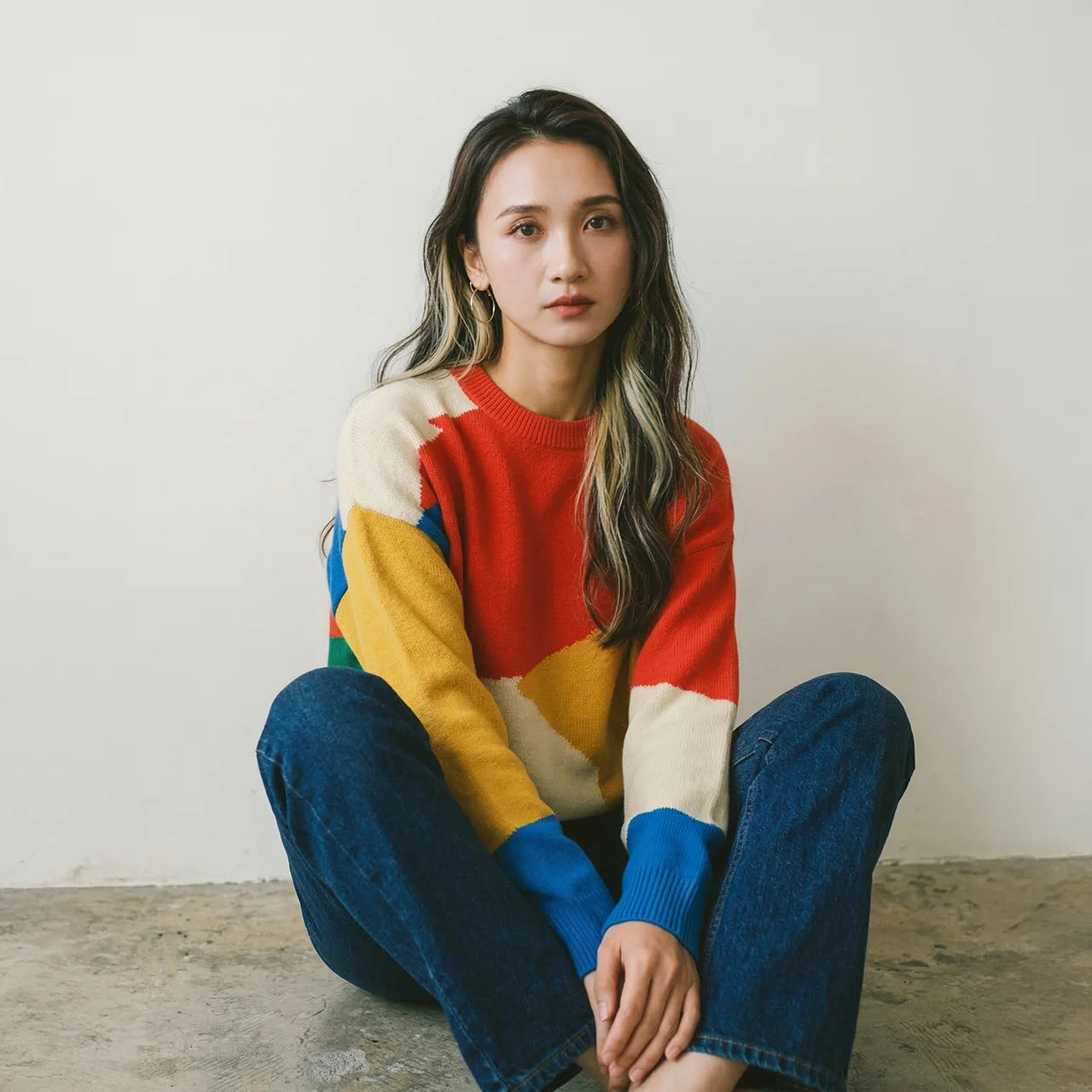 Woman seated on the floor against a plain white wall, wearing a colour-block knit jumper in red, yellow, blue, green, and cream with dark blue jeans.