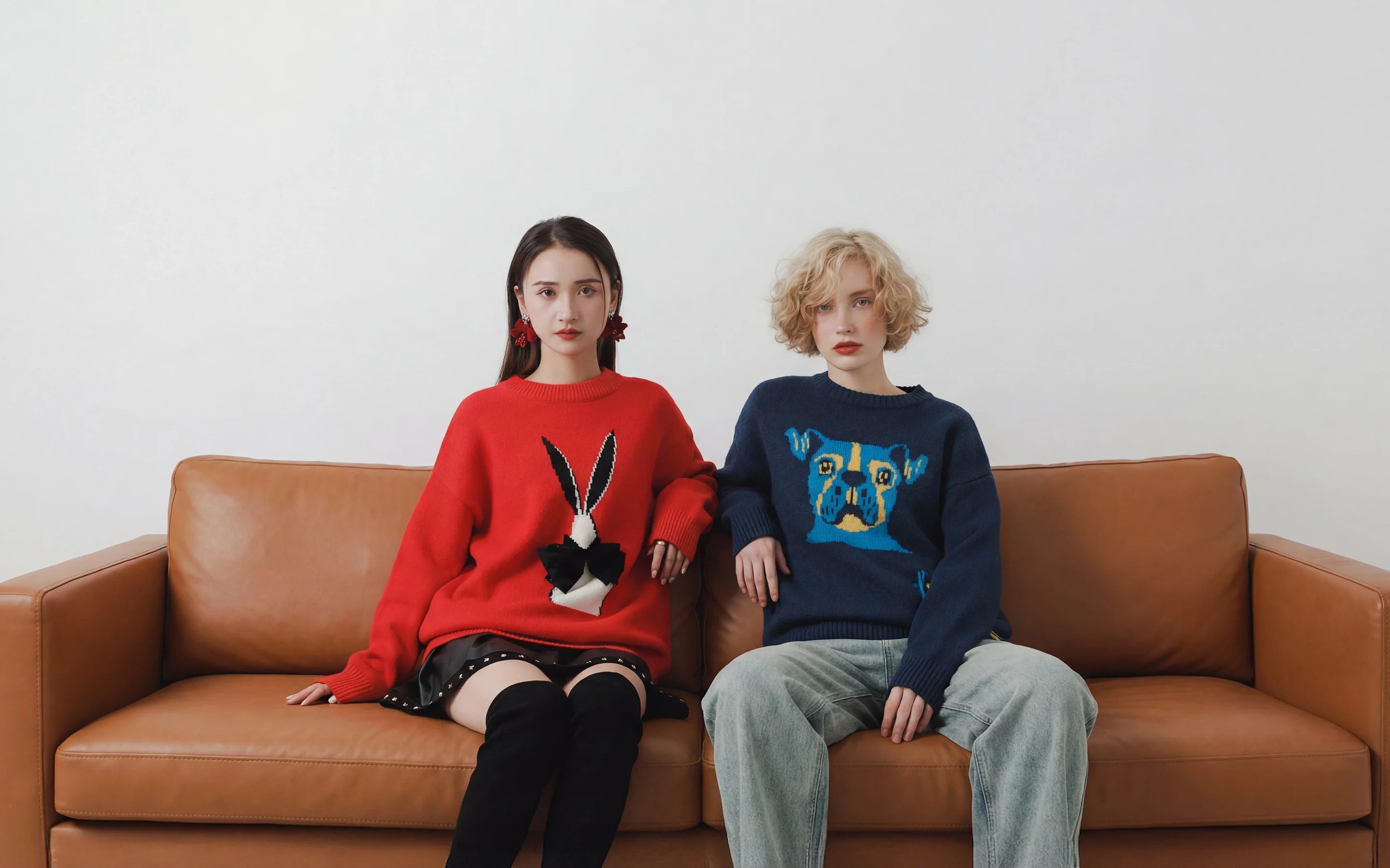 A wide homepage banner showing two women seated on a tan leather sofa against a pale wall, wearing oversized jacquard jumpers in red and navy with animal motifs.