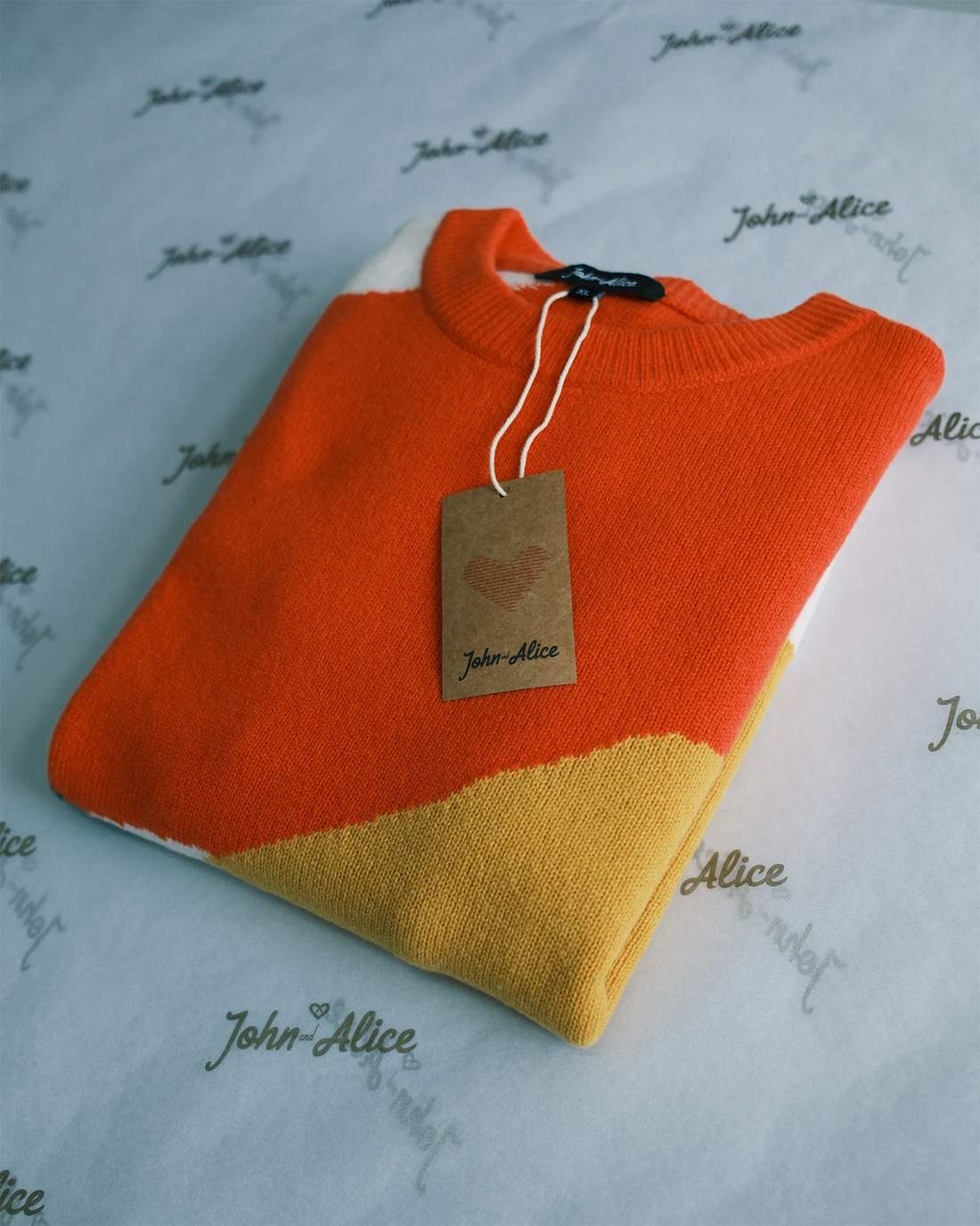 Folded orange and yellow knitwear with a John and Alice tag on branded tissue paper.