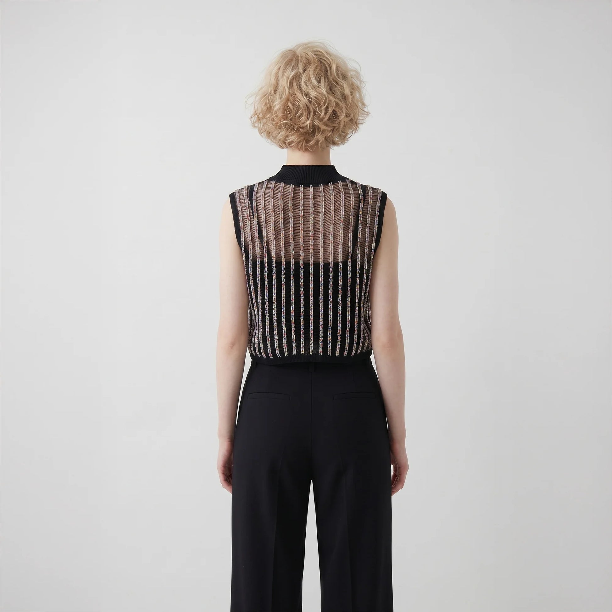 Back view of a model wearing a sleeveless cropped black mock neck crochet top with sheer upper panels and vertical textured line details, paired with black trousers against a plain light background.
