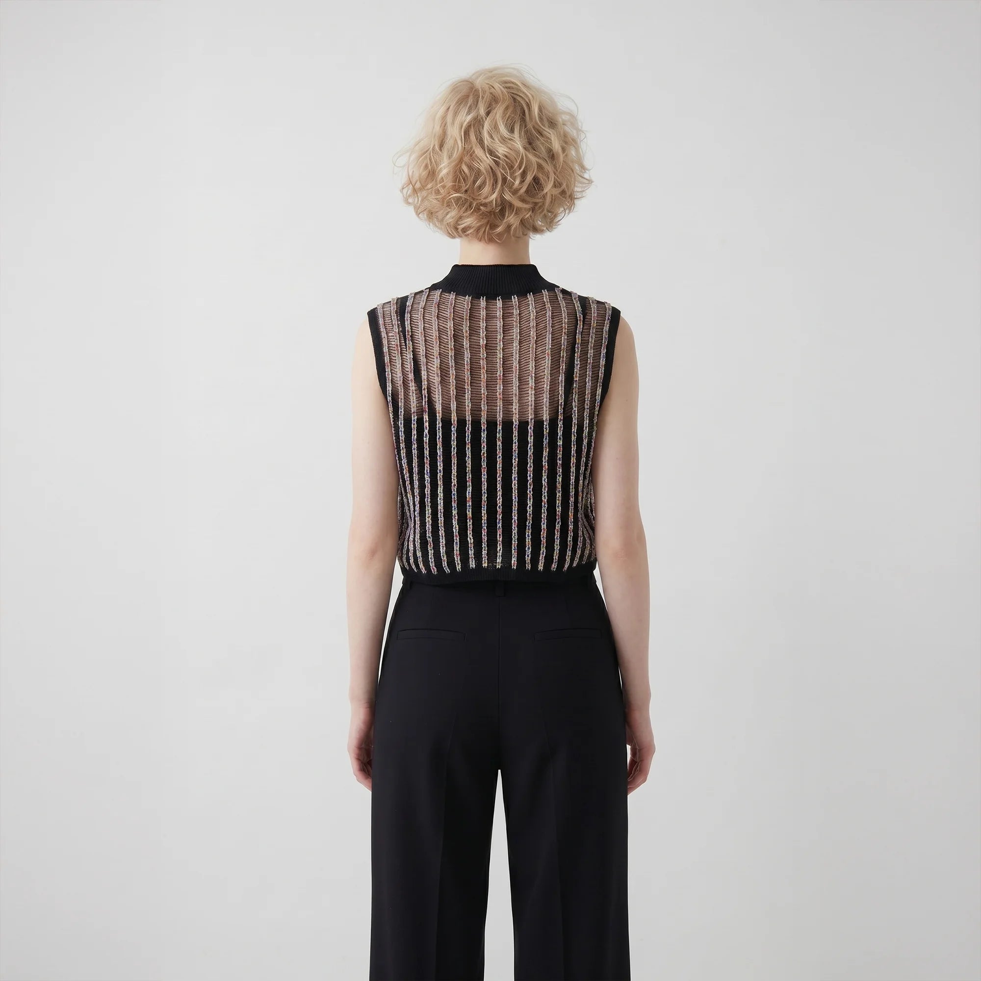 Back view of a model wearing a sleeveless cropped black mock neck crochet top with sheer upper panels and vertical textured line details, paired with black trousers against a plain light background.