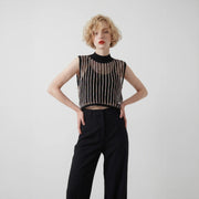 Blonde model wearing a sleeveless cropped black mock neck crochet top with vertical textured lines and high-waisted black trousers, shown from the front against a plain light background.