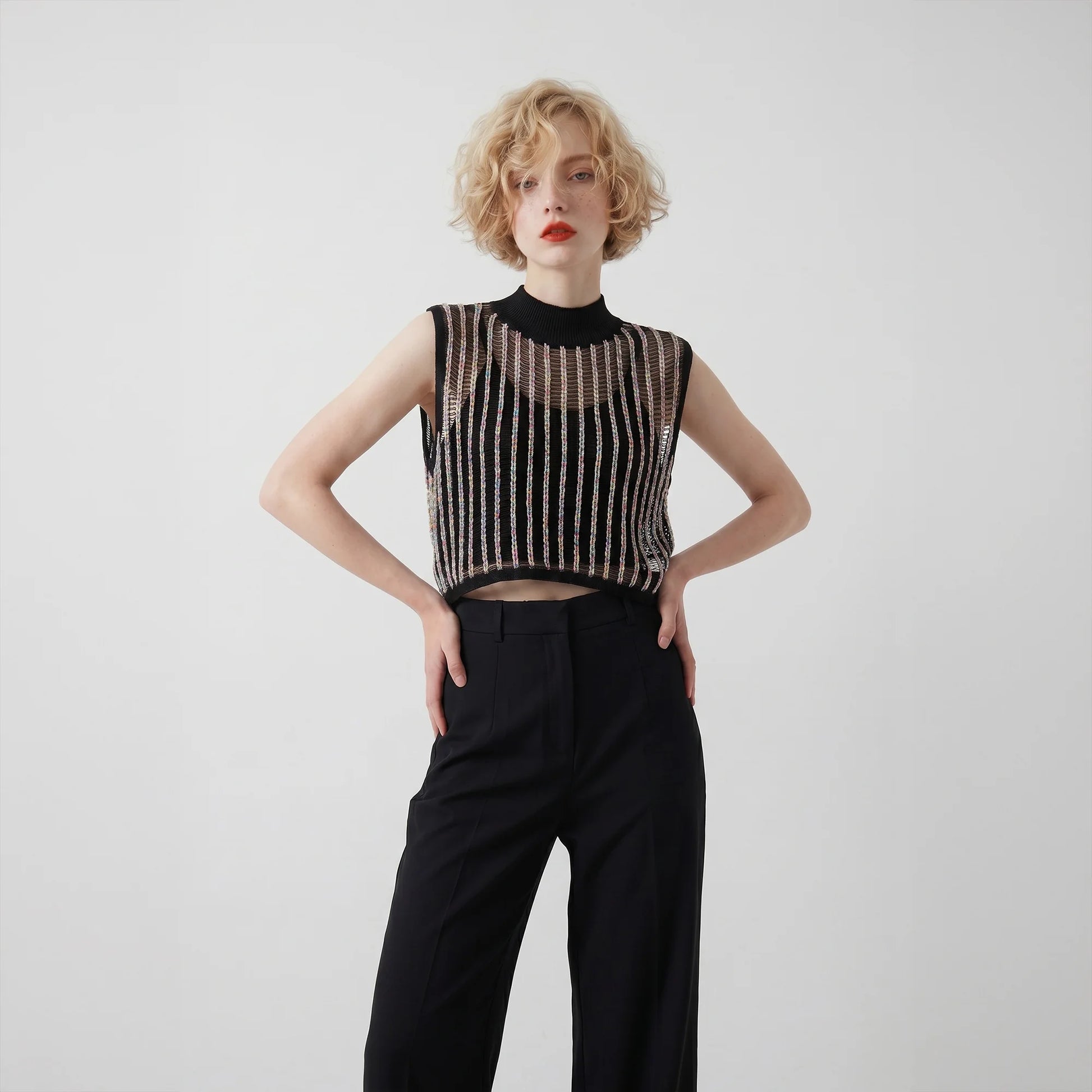Blonde model wearing a sleeveless cropped black mock neck crochet top with vertical textured lines and high-waisted black trousers, shown from the front against a plain light background.