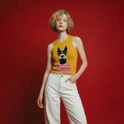 Model wearing a mustard yellow sleeveless knit tank with a dog intarsia motif and white jeans against a red background.