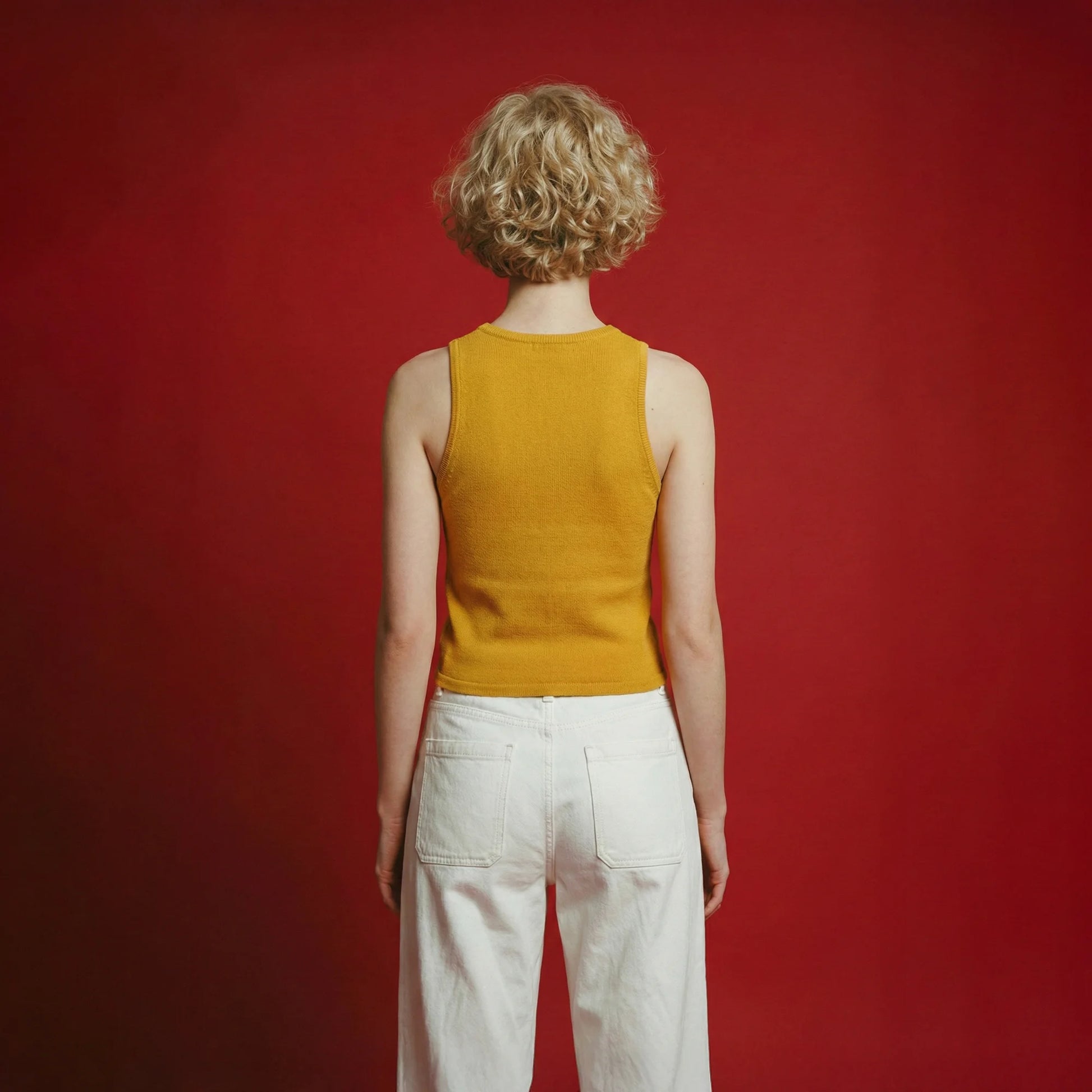 Back view of a model wearing a mustard yellow sleeveless knit tank and white jeans against a red background.