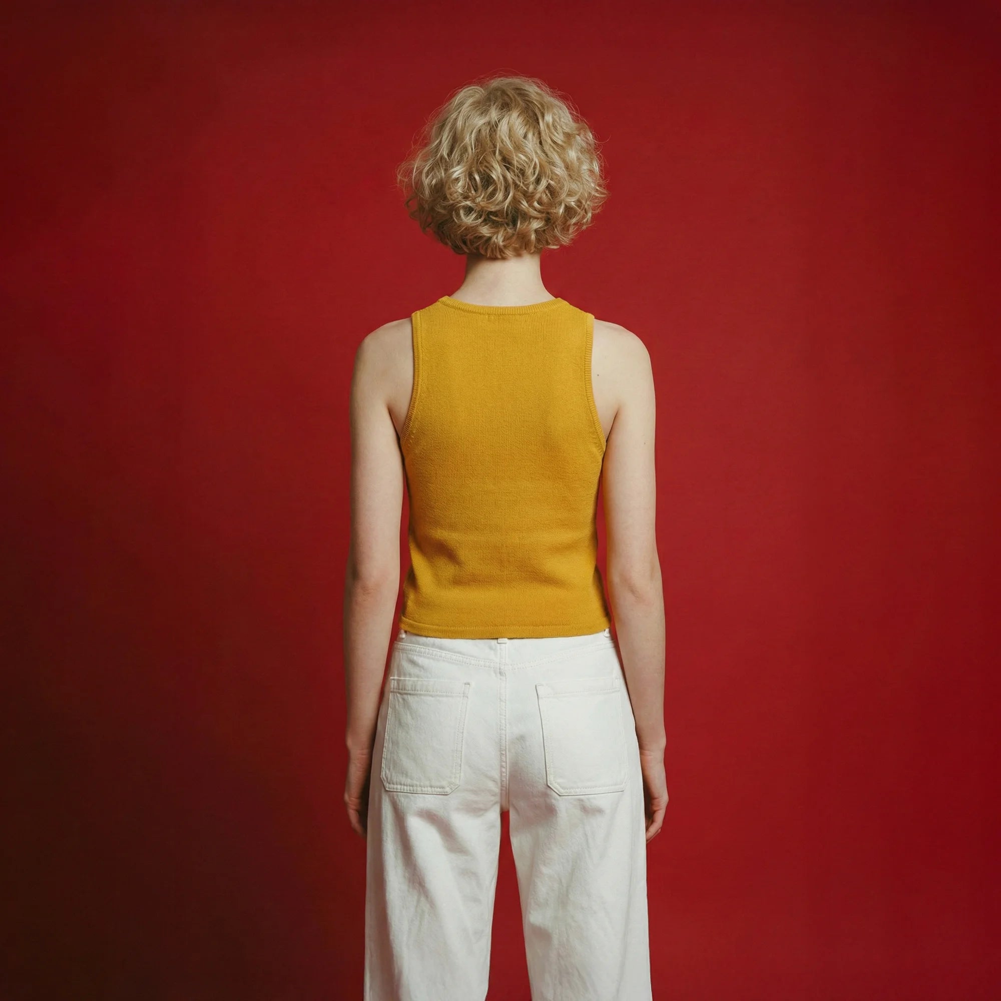 Back view of a model wearing a mustard yellow sleeveless knit tank and white jeans against a red background.
