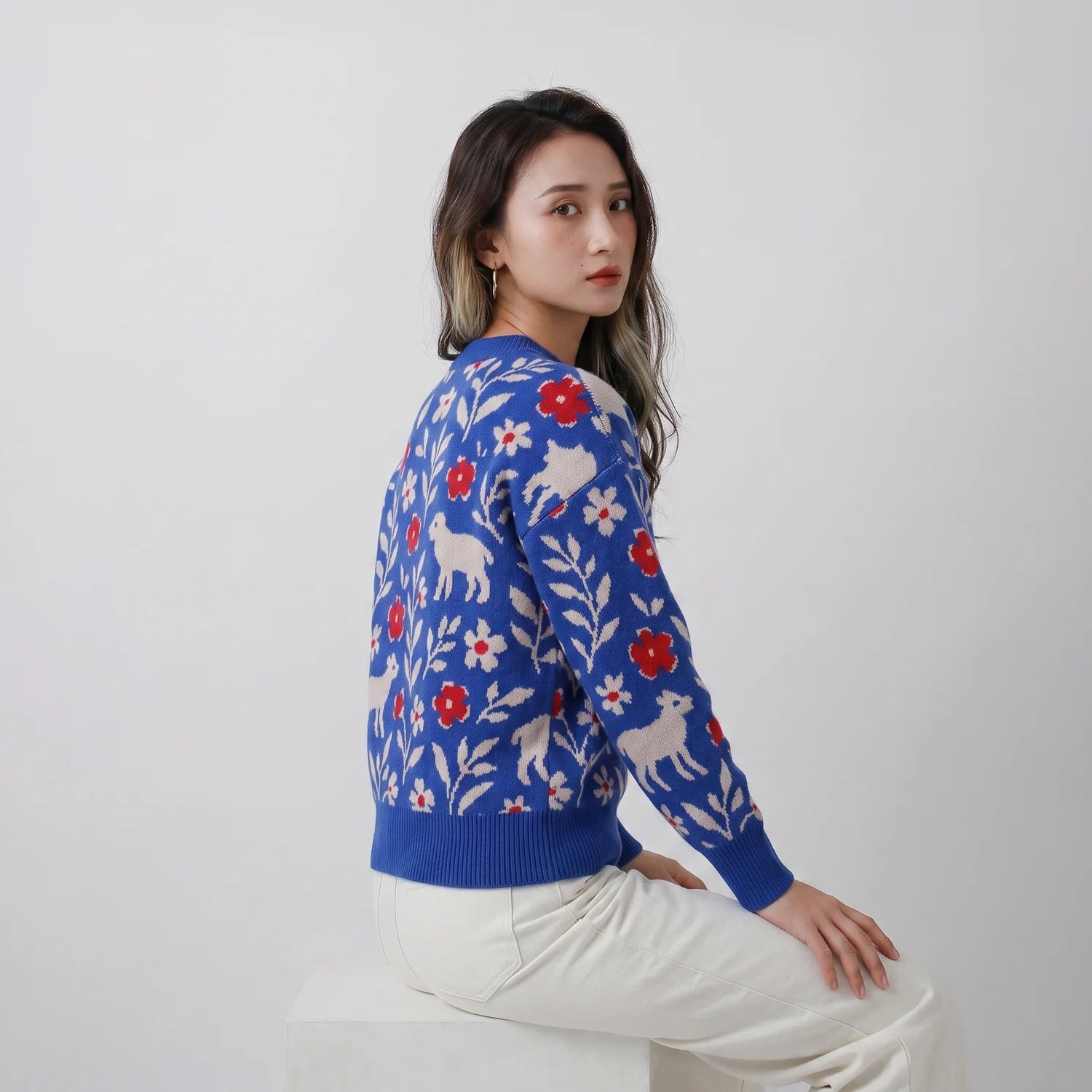 Seated model showing the back of a blue merino wool jumper with sheep and floral motifs.