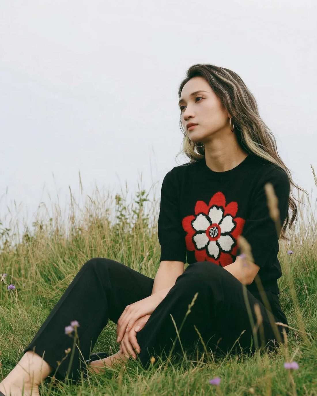 Collection card showing a woman seated in a grassy field wearing a black short-sleeve knit top with a large red and cream flower motif.