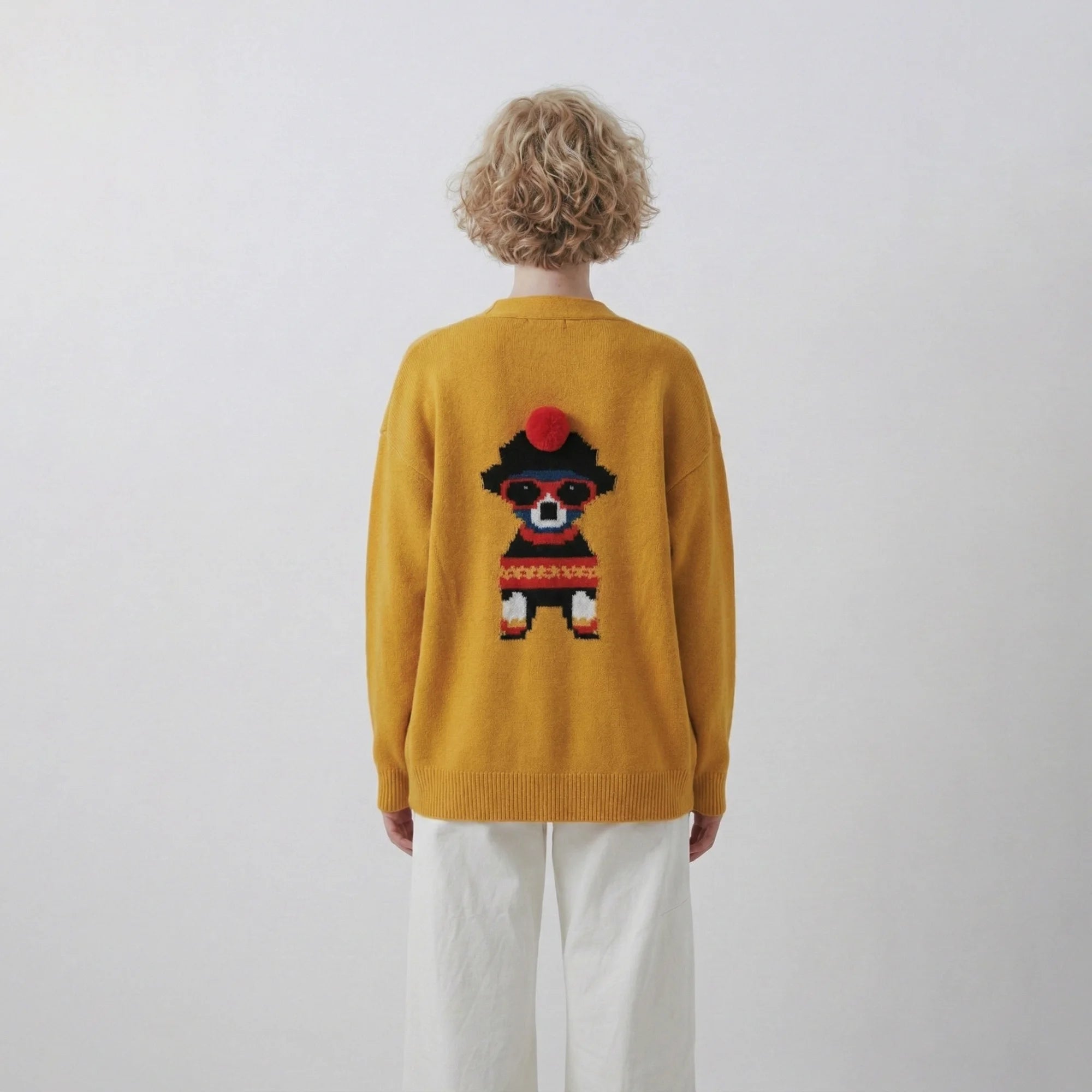 Model back view wearing a mustard yellow cardigan with a pixel dog jacquard and red pom-pom detail.