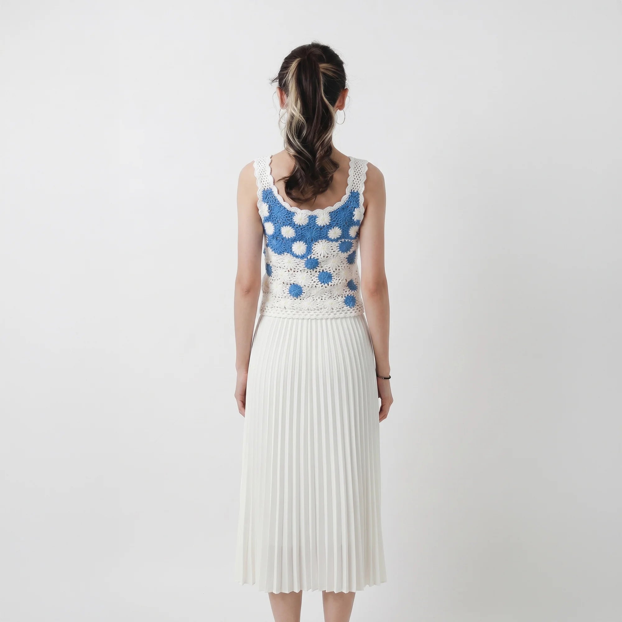Back view of model wearing a hand-crocheted fitted top in white and blue with floral motifs and small bead details, paired with a white pleated skirt.