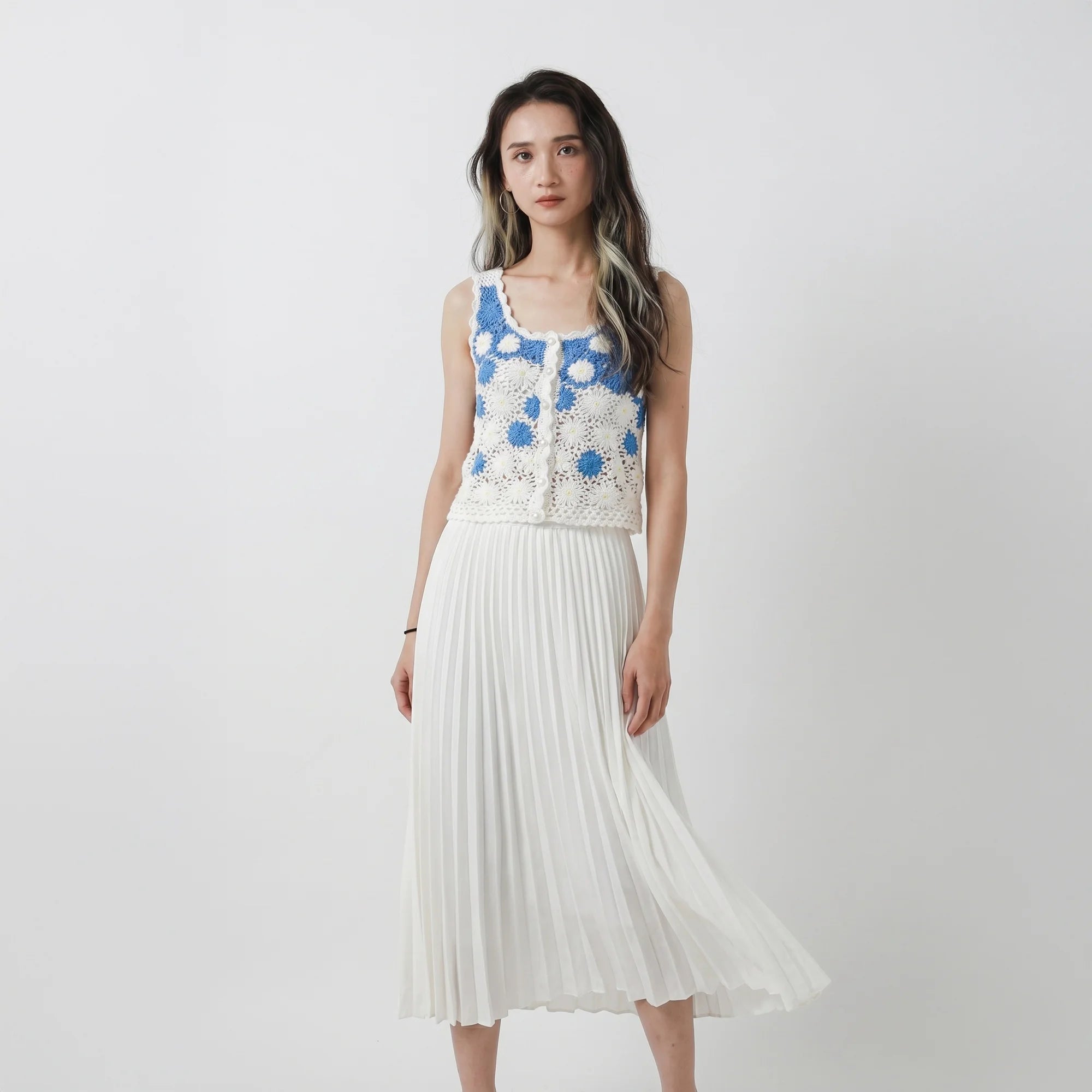 Model wearing a hand-crocheted fitted top in white and blue with floral motifs and small bead details, paired with a white pleated skirt.