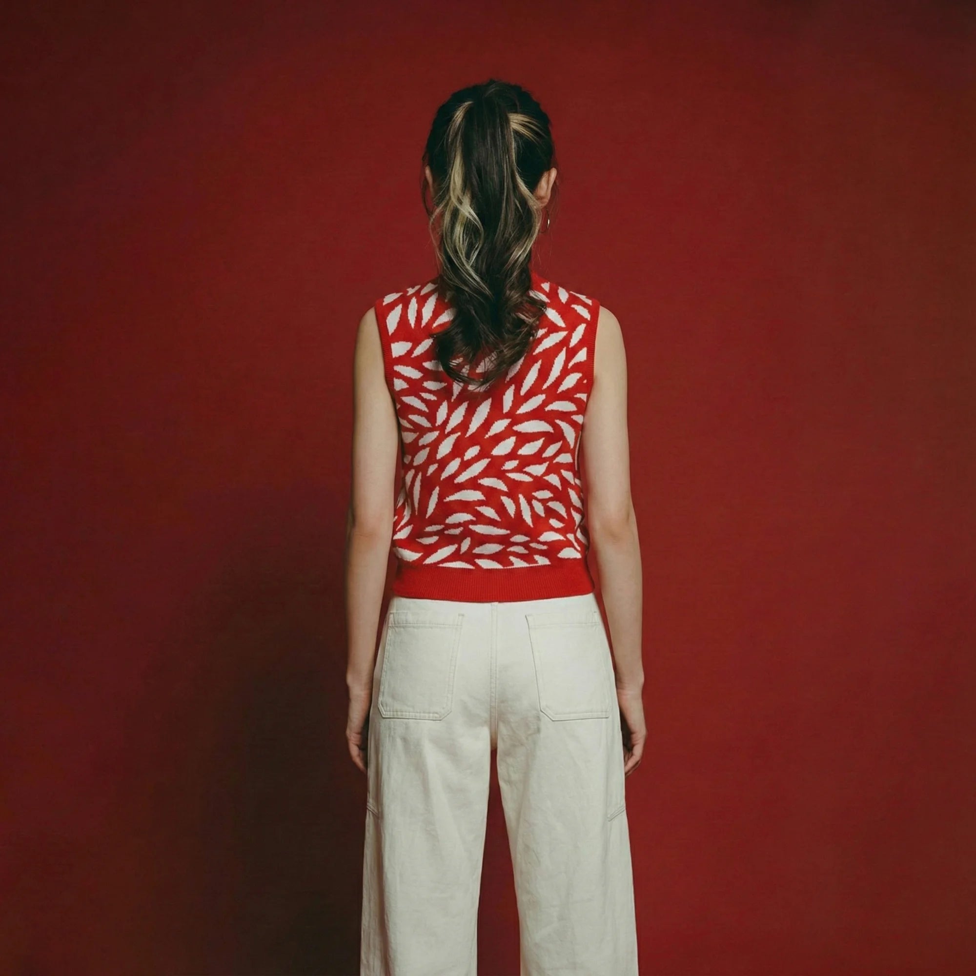 Back view of model in a red sleeveless leaf-pattern knit top and white trousers