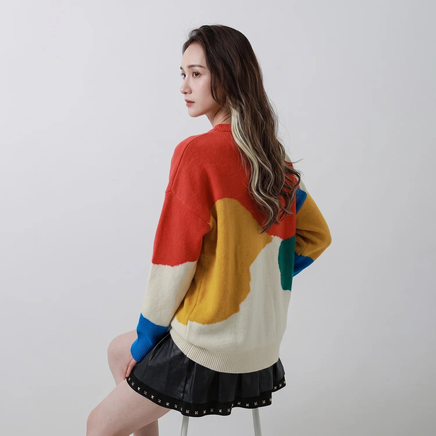 Seated back view of model wearing a coral multicolour merino wool jumper with abstract intarsia.