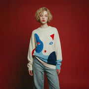 Model wearing a cream merino wool jumper with an oversized dropped-shoulder fit and a contrasting abstract cat intarsia on the front.