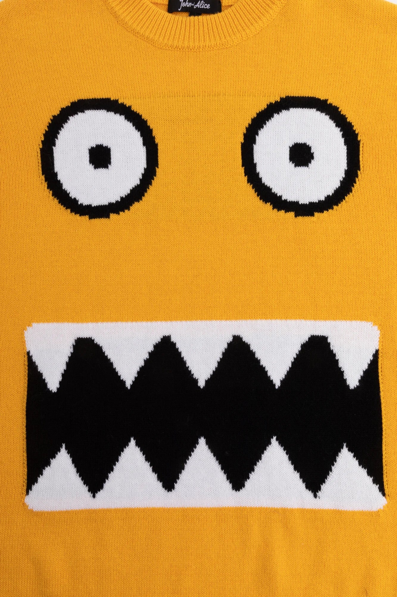 Close-up of the monster face jacquard with round eyes and a jagged mouth on the mustard yellow jumper.
