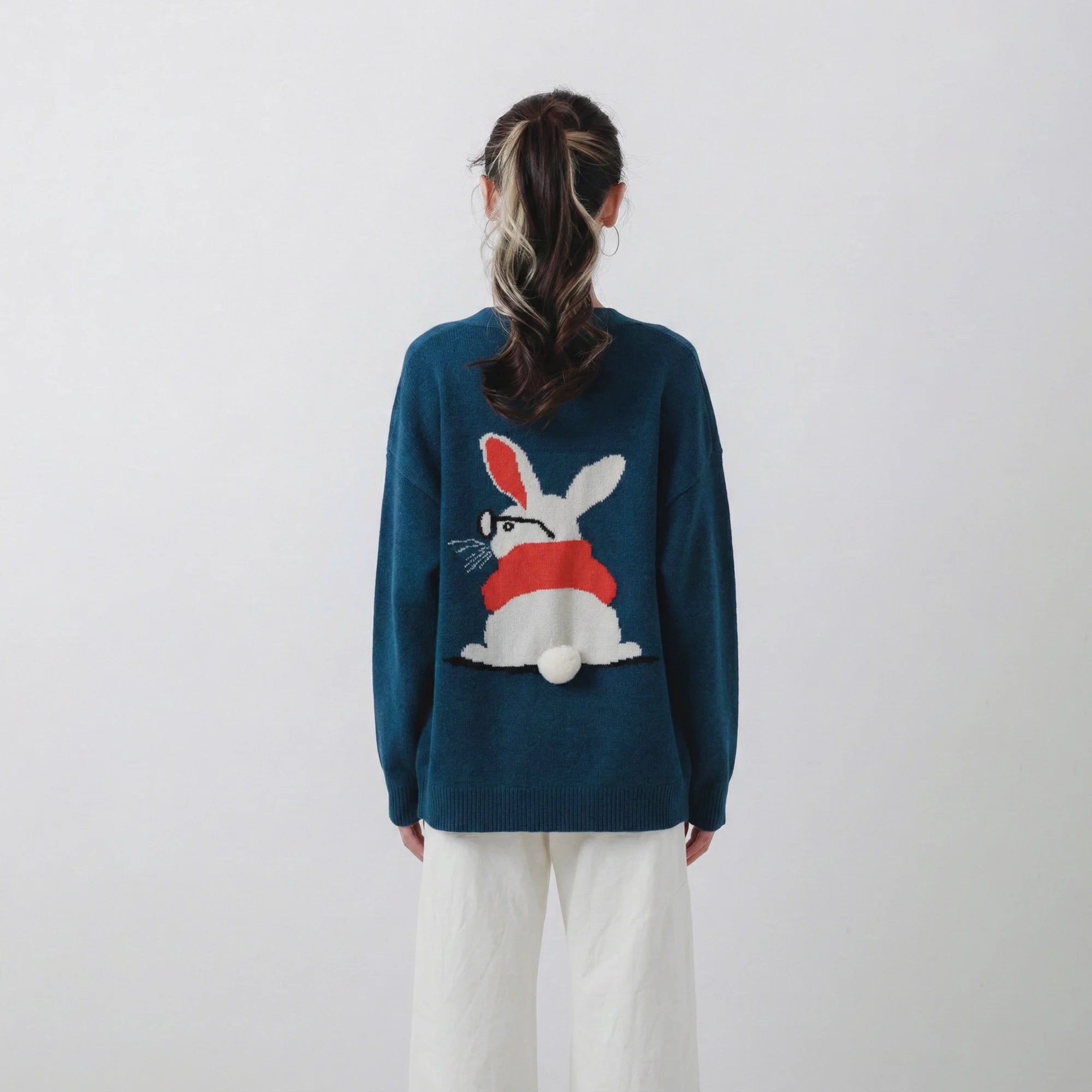 Back view of model wearing a teal blue merino wool cardigan with a rabbit intarsia and a pom-pom tail detail.