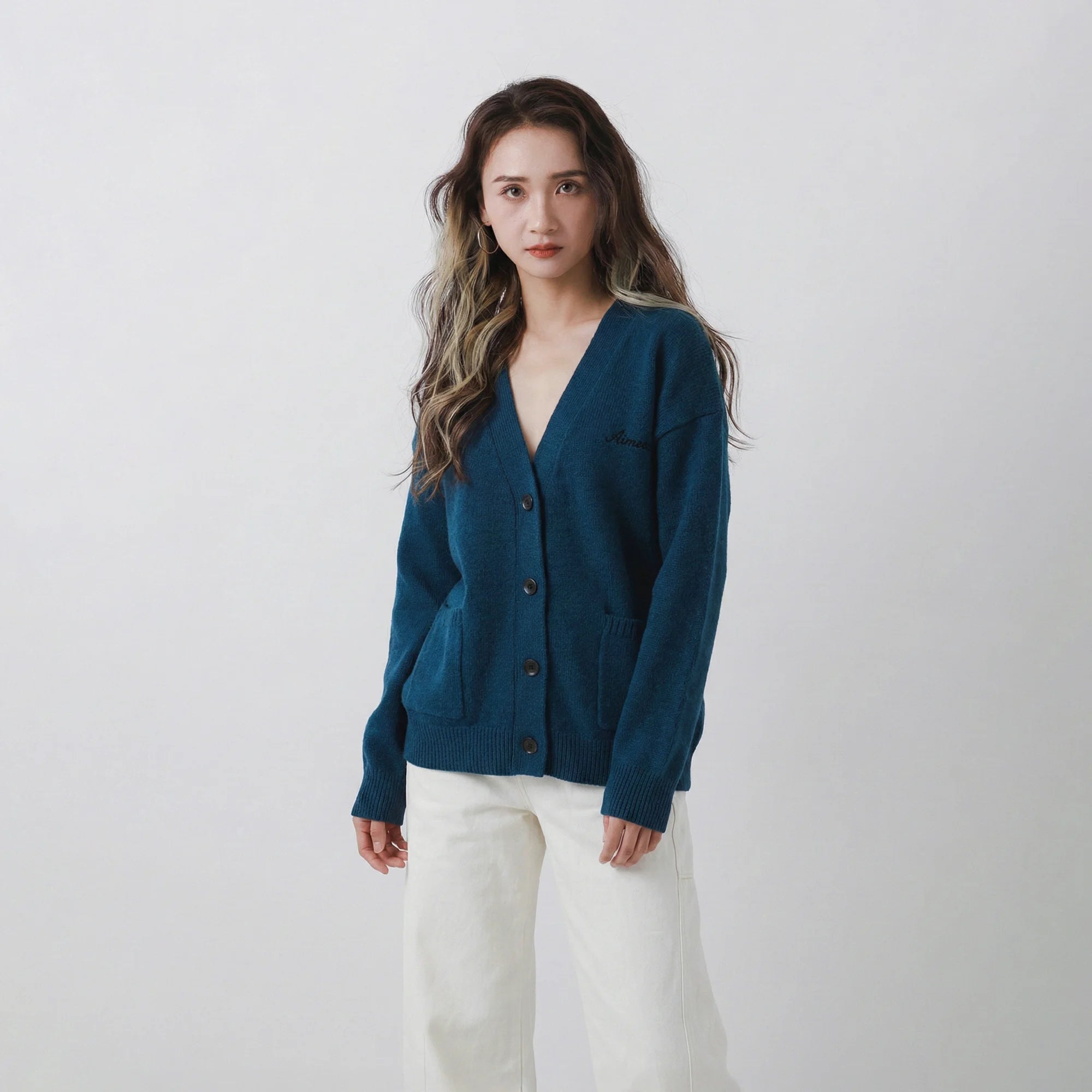 Model wearing a teal blue merino wool cardigan with a V-neck, patch pockets and John and Alice logo embroidery on the chest.